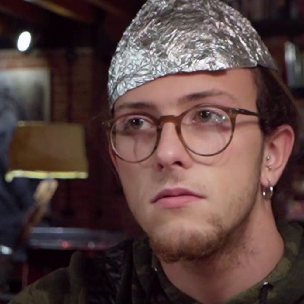 Filmstill aus dem Kurzfilm "Lie Detector". Ein junger Mann sitzt in einer WG Küche und trägt einen Hut aus Alufolie auf dem Kopf.