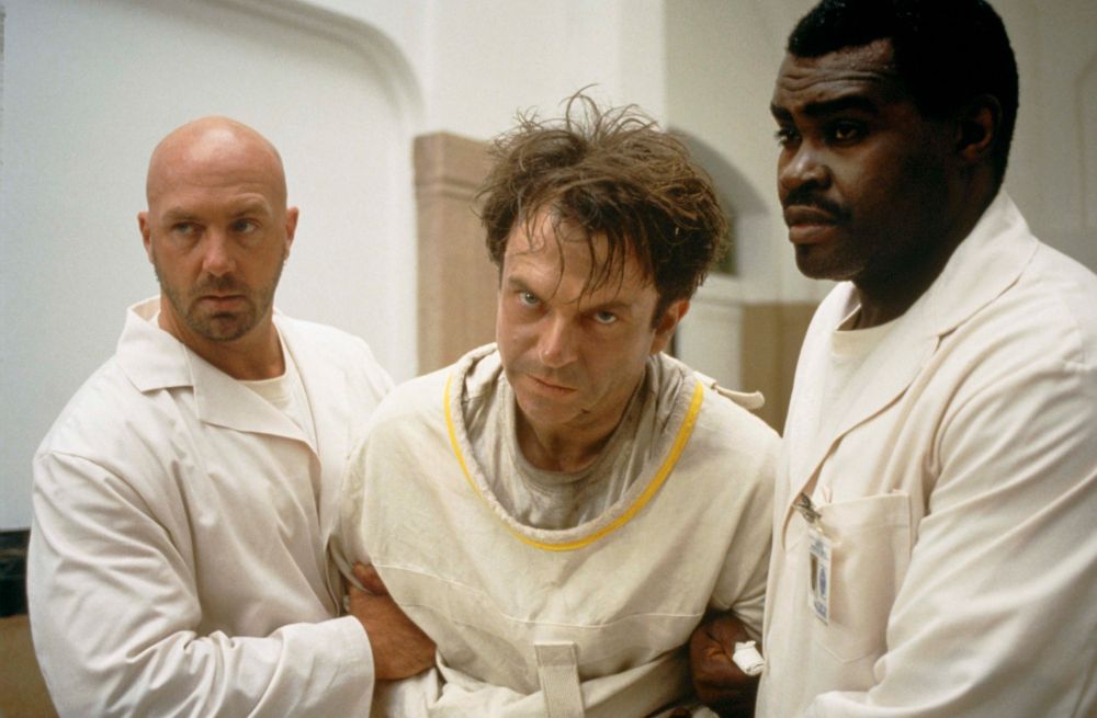 Sam Neill, circa 1990. In the Mouth of Madness and in a straitjacket with two men in white coats holding him by each arm.