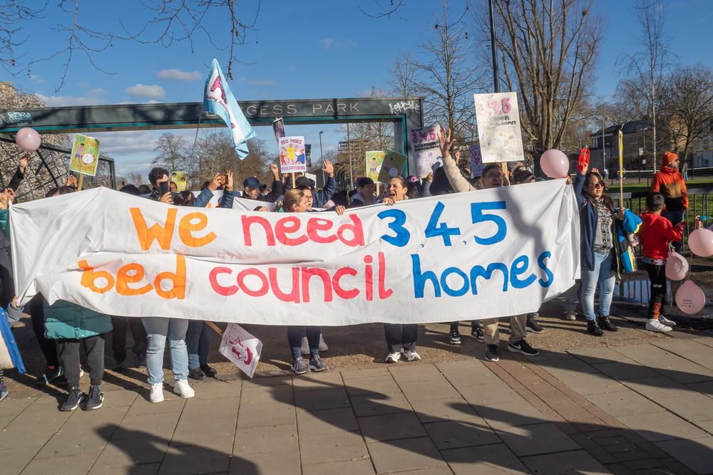 A large group of members from Housing Action Southwark and Lambeth at the entrance of Burgess Park holding a big banner reading 'We need 3, 4, 5 bed council homes'