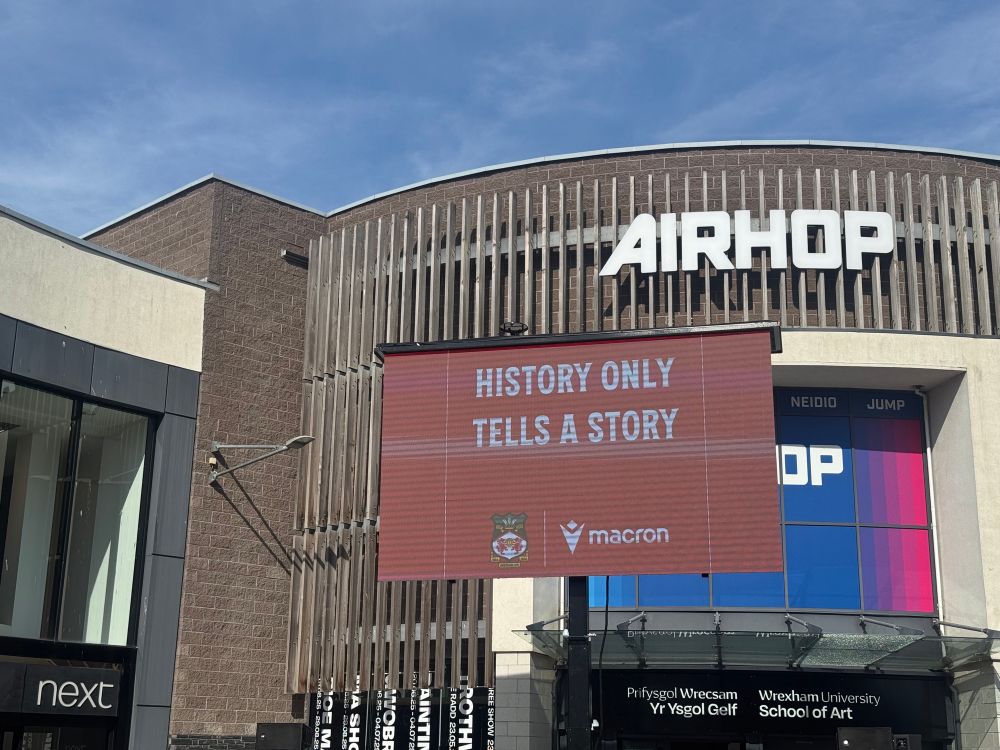 Big sign in Eagles meadow, Wrexham with the Wrexham AFC and Macron logos below the text “History only tells a story”