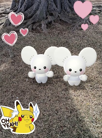 Tandemaus in Pokemon Go AR camera mode in front of a tree that has a decently large trunk and roots. The grass is being shaded by the tree. There's two different heart stickers placed on the top of the picture and an "OH YEAH!" Pikachu sticker on the bottom left.