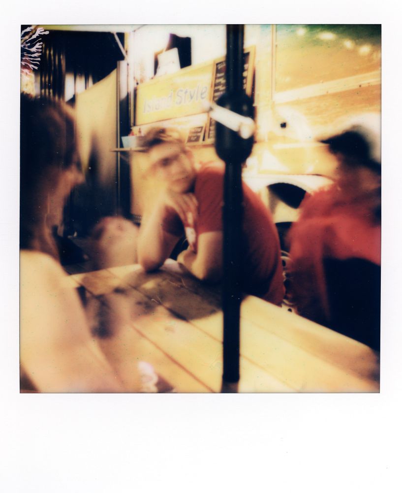 Color Polaroid of a man sitting at a picnic table. The photo is blurry and the man’s face is slightly distorted.