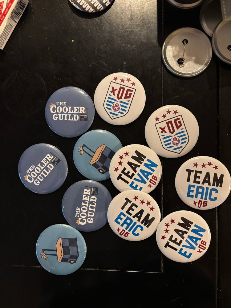 Pin-back buttons on a table featuring the logos of Expected Own Goals and The Cooler Guild. Some say “team evan” and “team eric” with xOG styling. 