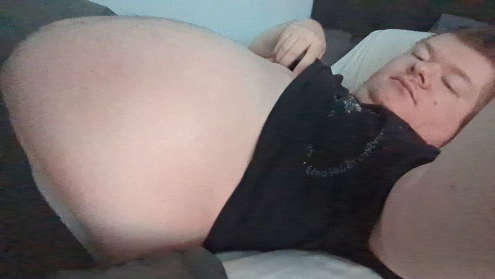 A selfie of Kit laying in bed. They're wearing black gym shorts and a black shirt, which has been pulled up to reveal their belly. Their face has a tired expression.