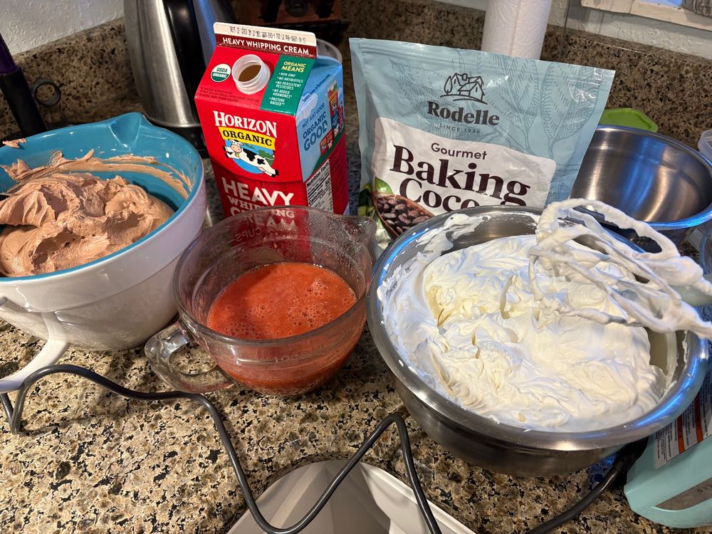 A mixing bowl full of chocolate mousse, one with strawberry puree, one with whipped cream. A carton of heavy whipping cream, and a package of baking cocoa in the background.