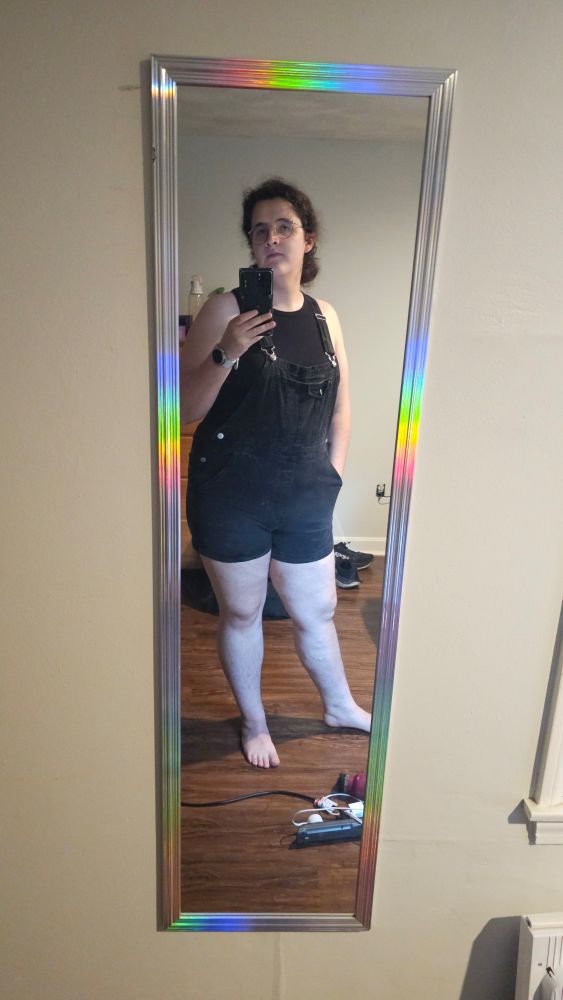 Sylvia in thrift overalls and a black tank top standing in front of a mirror