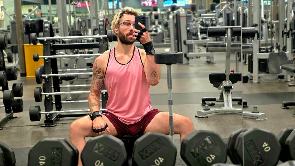 gym mirror selfie of a man spreading his legs on the bench wearing a pink tank top and maroon shorts and black lifting gloves 