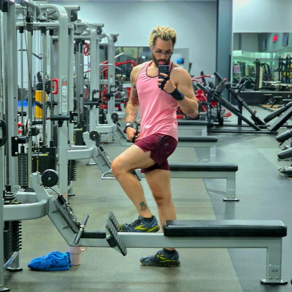 gym mirror selfie of a man in a pink tank top and maroon shorts 