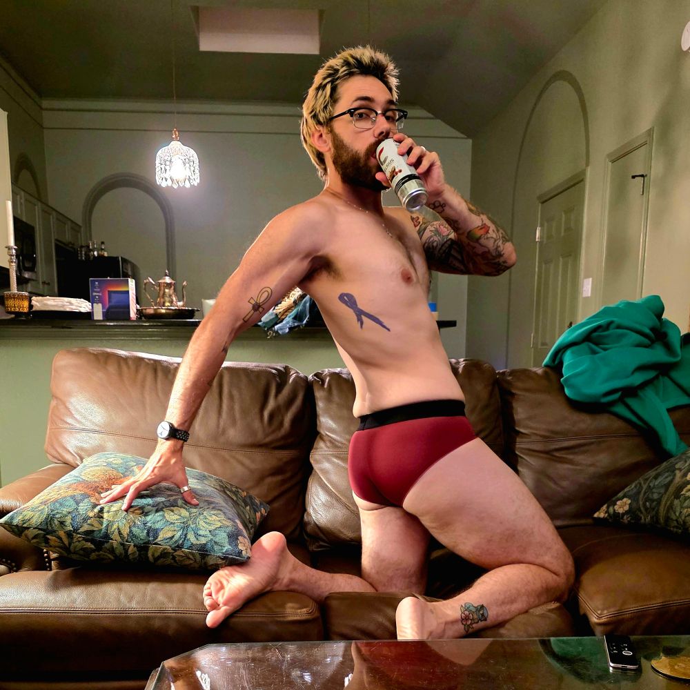 a man wearing only red briefs posing on the sofa showing off his ass while he sips a Celsius 