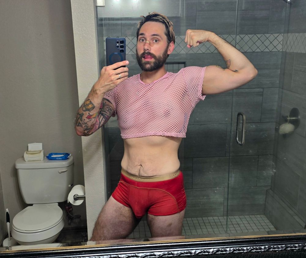 mirror selfie of a man flexing wearing a pink mesh crop to and red mesh underwear 