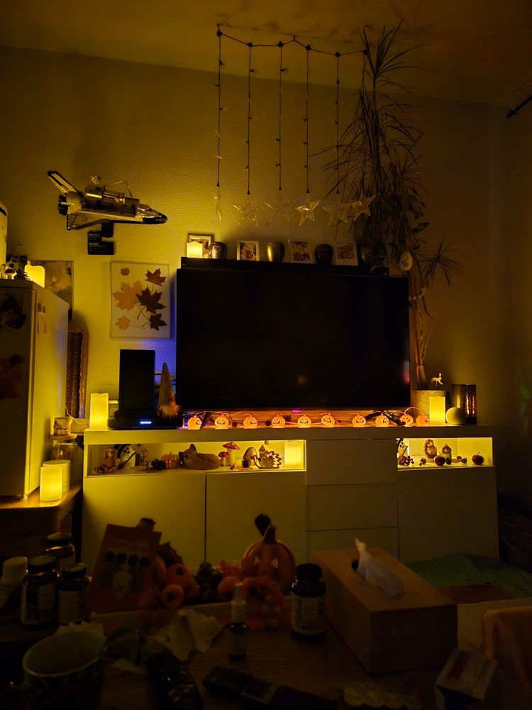 Das Bild zeigt eine dunkle Zimmerecke, die durch warmes, gelbliches Licht stimmungsvoll beleuchtet ist, was eine gemütliche Atmosphäre schafft, möglicherweise für den Herbst oder Halloween.
​Zentrale Einrichtung: Im Zentrum steht ein großer, dunkler Fernseher auf einem weißen Schrank oder Lowboard.
​Beleuchtung und Dekoration:
​Das Lowboard wird von unten durch eine indirekte Beleuchtung in warmem Gelb beleuchtet.
​Auf der Oberseite des Lowboards, vor dem Fernseher, verläuft eine Lichterkette mit Halloween-Kürbissen.
​Im oberen Bereich hängen mehrere Stränge von Sternen-Lichterketten herab.
​Mehrere Kerzen (scheinbar elektrische oder LED-Kerzen) sind im Raum verteilt, unter anderem auf dem Lowboard und links neben dem Schrank, was zur gelben