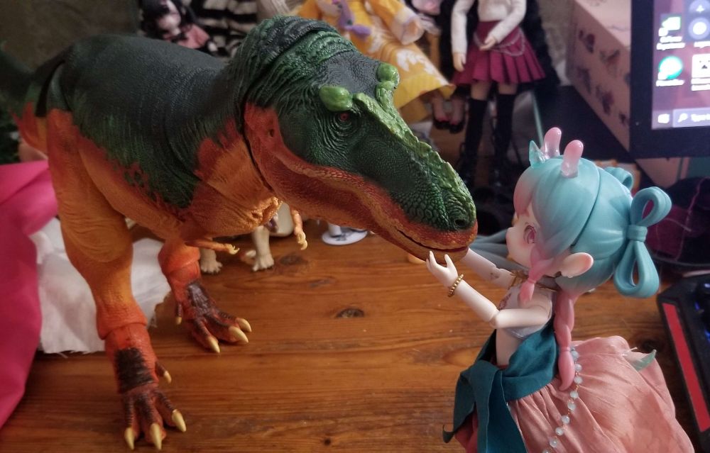 a small deer girl bjd is posed gently holding the snout of a much larger and more realistic tyrannosaurus figure.