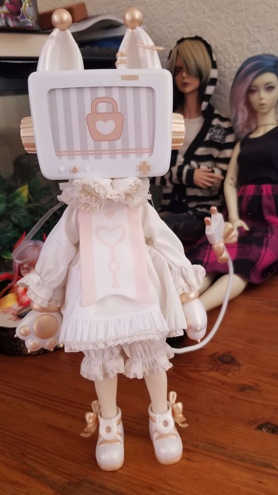 A medium sized ball jointed doll. The doll has a television for a head with a pair of cat ears on top. The doll is wearing a ruffly white and pink dress, and has cat paws for hands and an electrical plug for a tail.