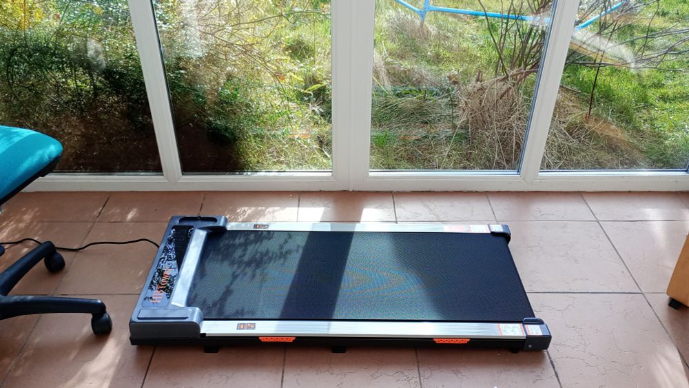 A walking pad by conservatory windows. It's a smaller, lighter weight, slower version of a treadmill. 