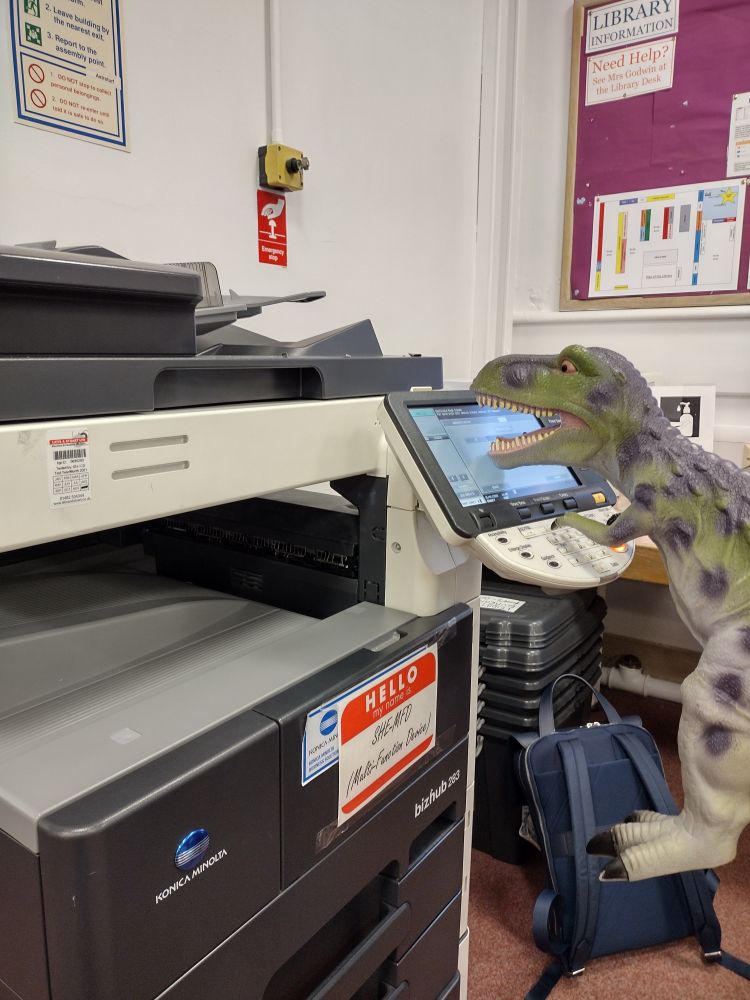 A (toy) t-rex using a large grey photocopier. He's struggling to press the buttons becaus eof his tiny arms. 