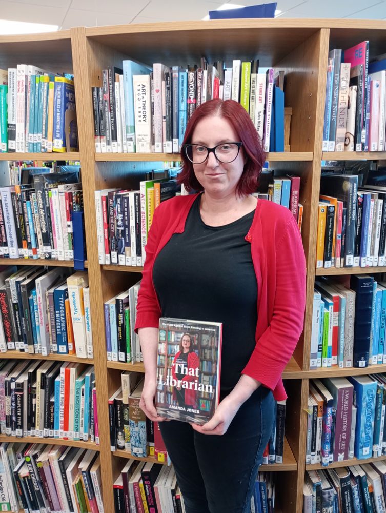 A woman with glasses and shoulder length red hair, wearing a black top, black trousers and a red cardigan stands in front of a bookcase, holding the book That Librarian which features it's author, Amanda Jones, on the cover. the cover photo shows Amanda standing in front of a bookcase, she is wearing glasses, has shoulder legnth brown hair and is wearing a black Freadom t-shirt, black trousers and a red cardigan. 