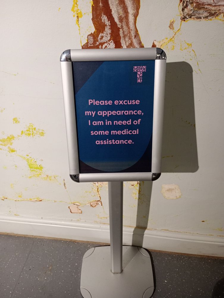 Please excuse my appearance, I am in need of some medical assistance. A sign from the Thackray Museum, in front of a water damaged wall.