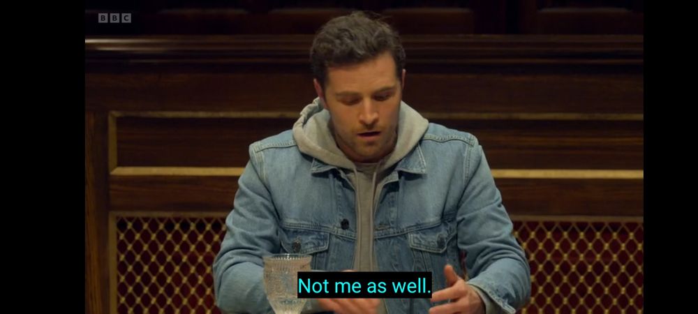 Alexander, from Traitors S3, realising he has misspelt Minah's name. He's sitting at the round table, a dark brown room, wearing a grey hoodie and denim jacket and has a look of mortification on his face, with the subtitle 'not me as well'.