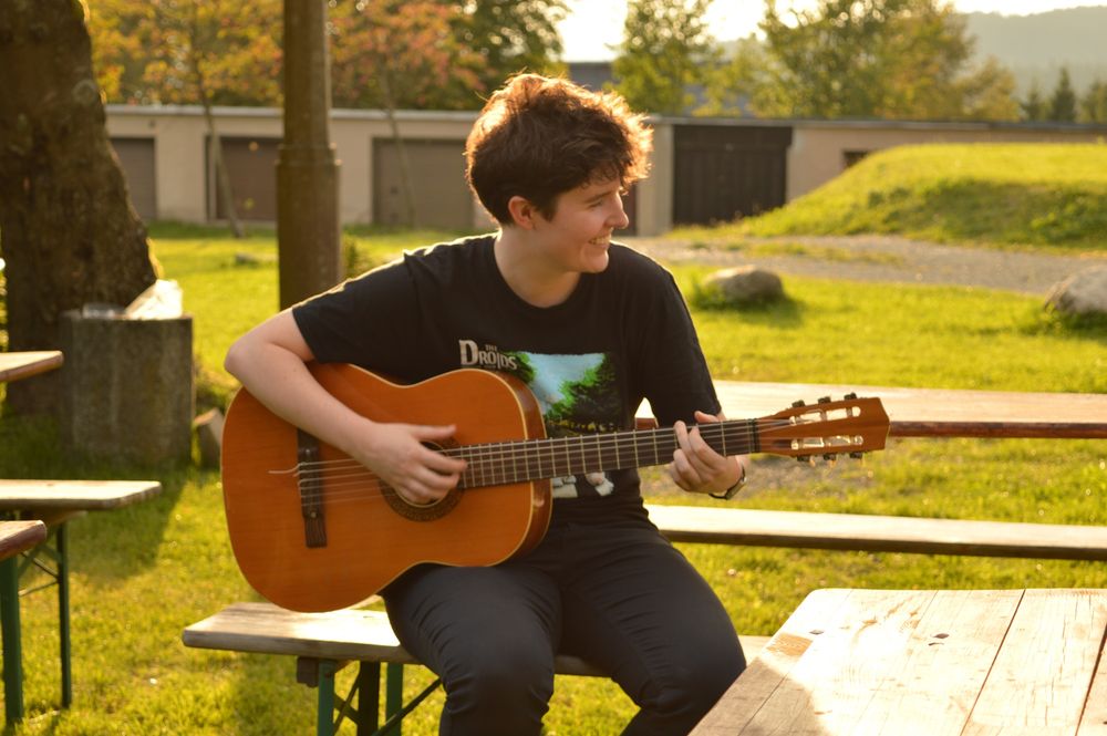 A happy guitarist sitting on a picnic bench in the summer sunshine. They are in their late 20s, white, with short brown hair and are smiling with their eyes closed. They’re playing an orange classical guitar and wearing a black t-shirt that shows a Star Wars parody of the Beatles’ Abbey Road album, where the Beatles are droids 