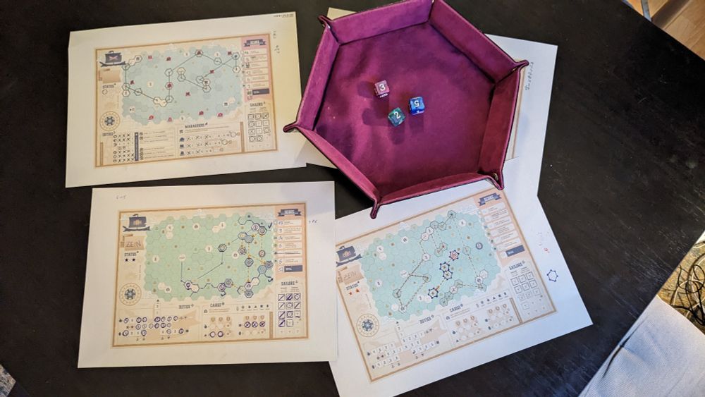 Photo of three printed sea maps with lines drawn on them marking the journeys, and other fields filled in for resources and such. There's a dice tray with three dice beside it.