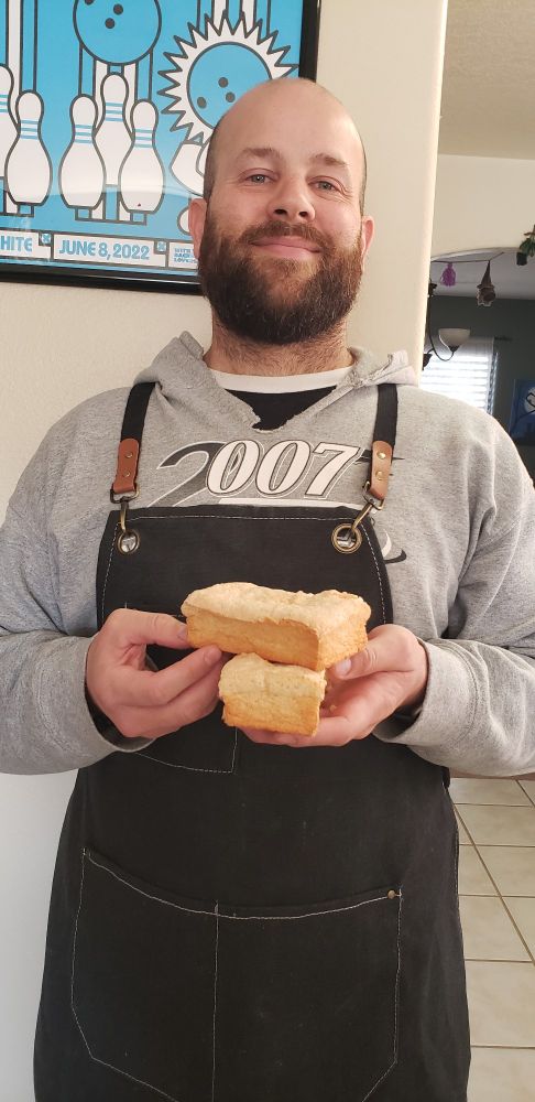 A man with a grey hoodie and black apron holding the homemade Gluten Free Angel Food Cake he made. 