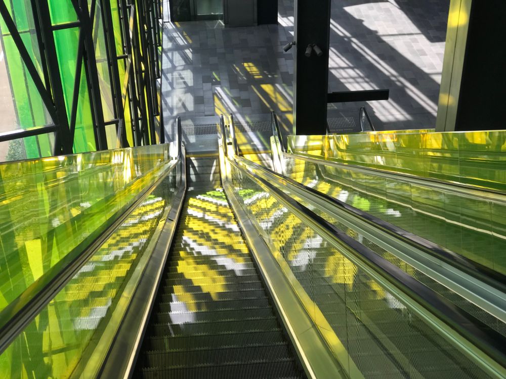 A view down an escalator towards ground level, with stripes of white, yellow, and green light caused by colored windows on the left