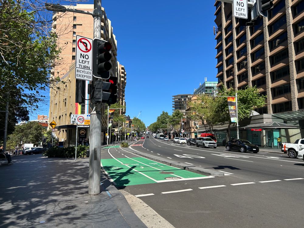 Looking along a wide road with a bidirectional cycleway on the left of the road, where the lane going towards the viewpoint splits into 2 lanes, 1 for going straight and 1 for turning right