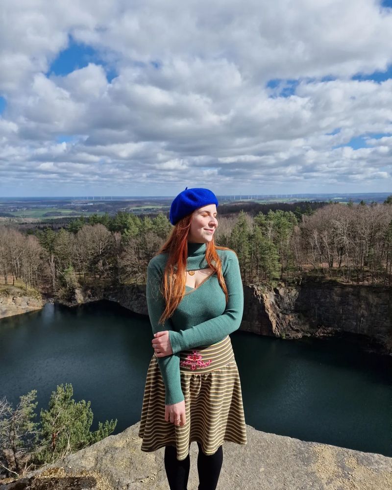 Ich stehe auf steinigen Untergrund, im Hintergrund sieht man einen Steinbruch. Das Foto wurde von jemand anderes aufgenommen. Ich habe die Augen geschlossen Gesicht richtung Sonne. Ich trage einen gestreiften Rock und ein langärmliges Oberteil