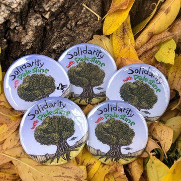 Image of several circular pin buttons, spread out over yellow leaves. Blue buttons have the same design. 
Design features an olive tree in the shape of a fist. Above this tree, text reads "Solidarity with Palestine".