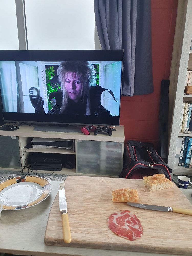 A photo of a TV showing David Bowie as Jareth in Labyrinth, with a mostly-eaten platter of homemade focaccia with hummus and charcuterie