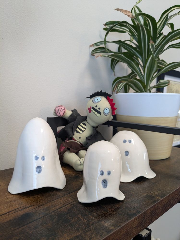 Three white ceramic ghosts of various shapes and sizes sit on a shelf. They have various "woooooo" spooky mouths. In the back is a plush zombie with a brain in its hand and blood dripping from its mouth.