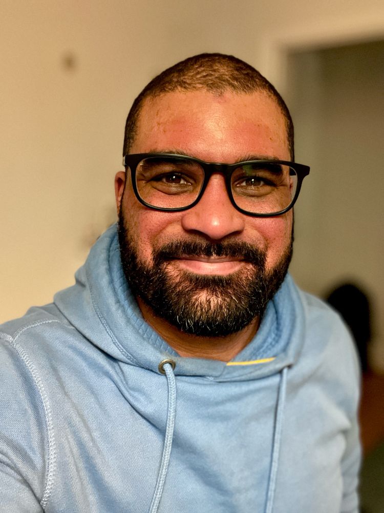 A selfie of Steve Gilbert in a baby blue hoodie. Steve has a well groomed black beard and moustache, short black hair and black glasses. 