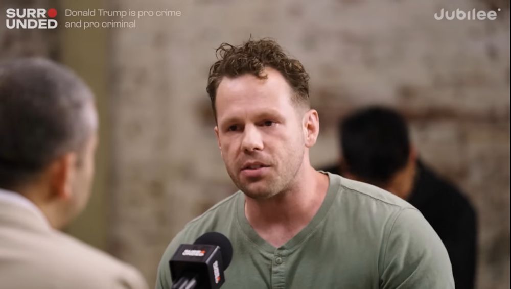 A white man with a fade haircut and brown curls on top wears an olive henley top.