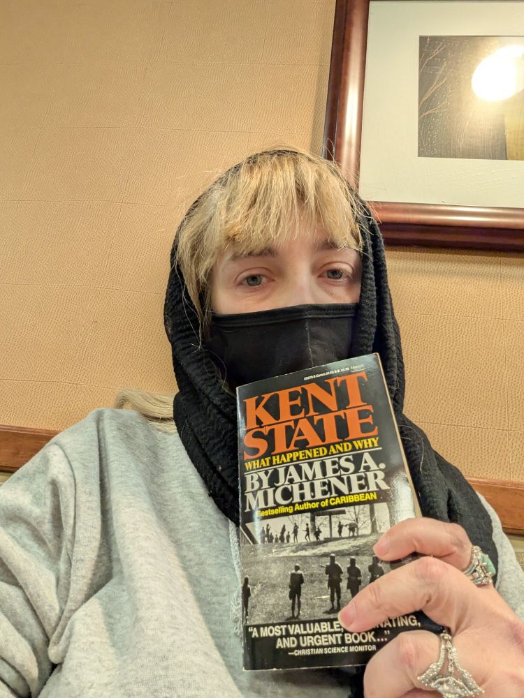 Woman in a mask holding 'Kent State what happened and why'd by James A. Michener