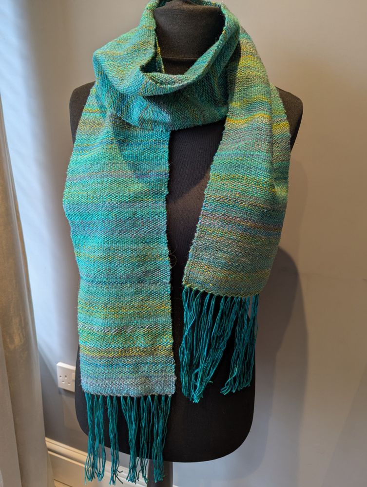 Hedy, a headless and armless black mannequin, sports a woven scarf in shades of teal and green. The warp is a slightly darker shade of teal.