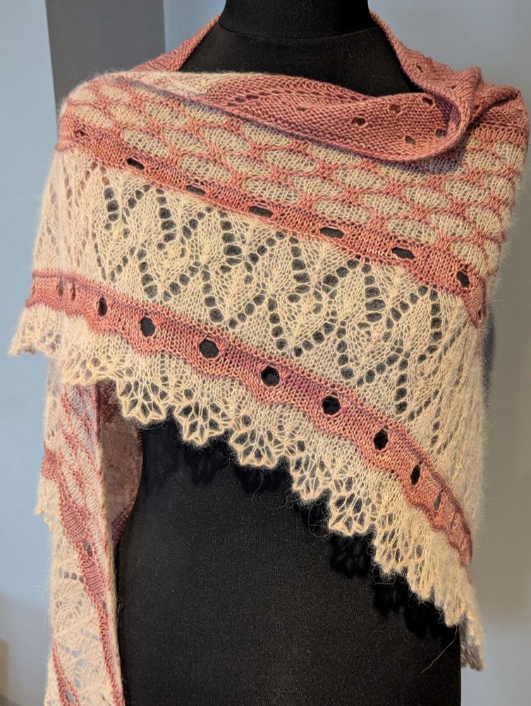 A better image of the lace on a shawl with a soft pinkish body and creamy peach lace edging on a headless mannequin against a slate grey background. The lace is an alpaca blend that fluffs into the stitches. It is perhaps softer to the touch than it looks, and it seems very squishy.