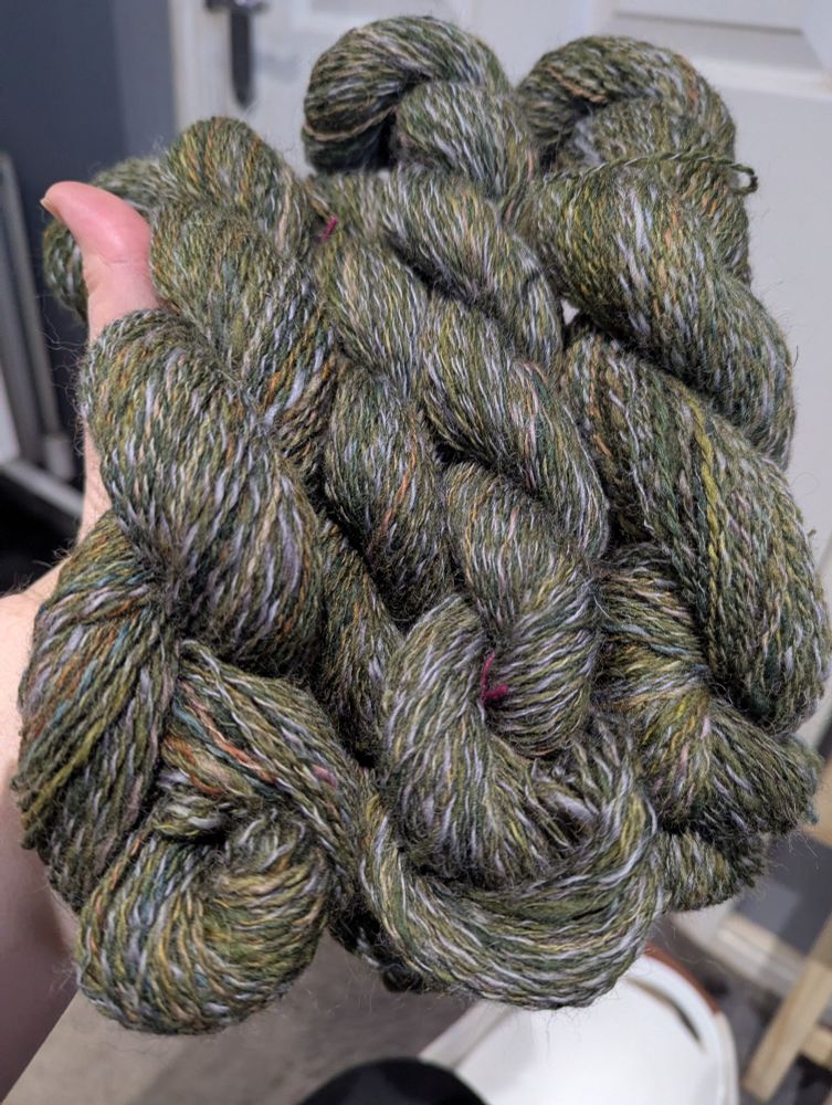 A woman's hand holds for smallish hanks of mottled green Shetland wool plied with a variegated grey, green, and brown wool/alpaca blend.