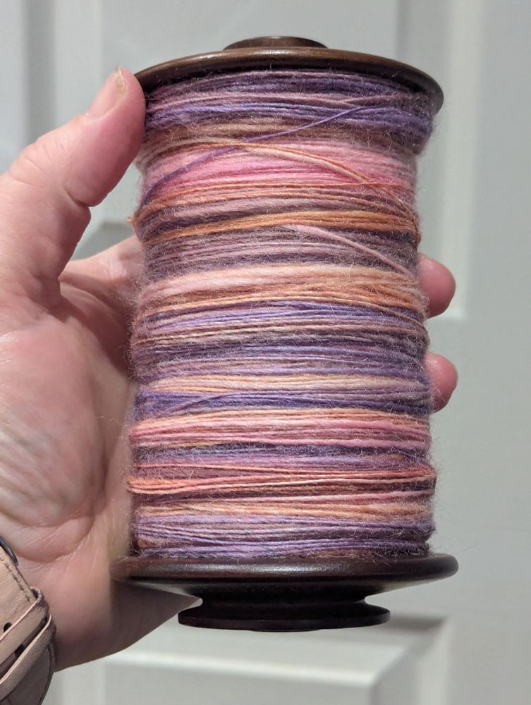 A spinning wheel bobbin filled to the brim with fairly evenly spun yarn in shades of pink, peach, and purple.