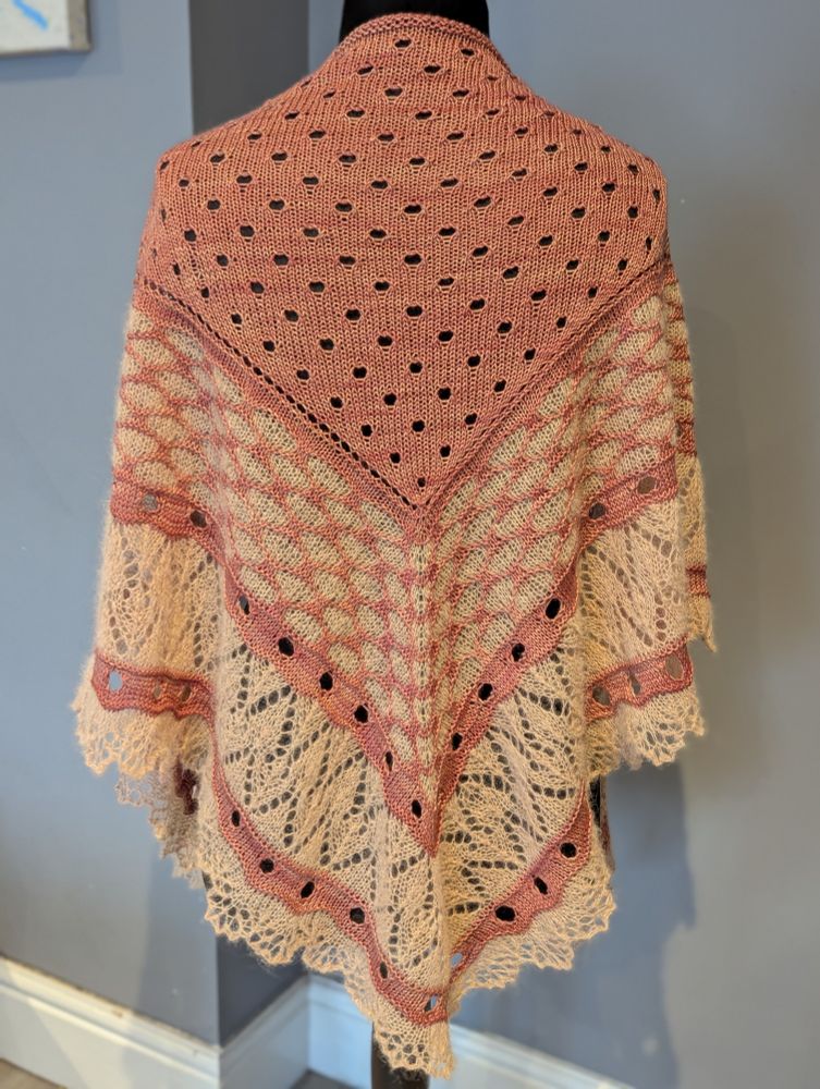 A shawl with a soft pinkish body and creamy peach lace edging on a headless mannequin against a slate grey background. It is perhaps softer to the touch than it looks, and it seems very squishy.