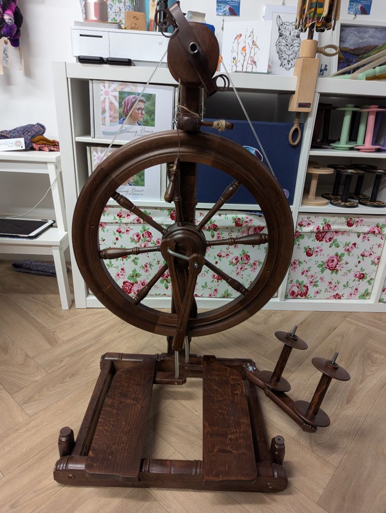 A walnut spinning wheel sits in front of a white wall and white shelves. There are floral fabric bins in the lowest cubbies of the shelf.