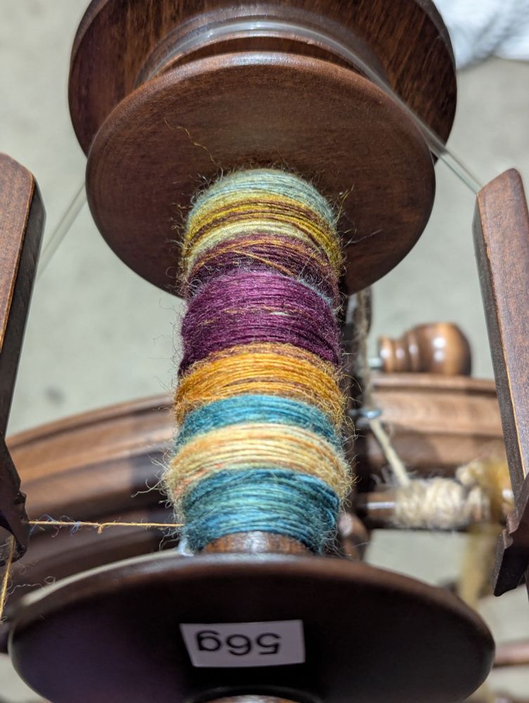 A multicoloured bobbin of dark reds, bright teal, rusty orange, and pale teal is mounted on a walnut spinning wheel. 