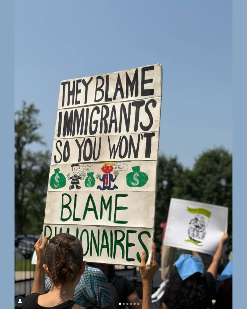 A protester holds a handmade sign that reads: “THEY BLAME IMMIGRANTS SO YOU WON’T BLAME BILLIONAIRES.” The sign includes cartoon drawings of a rich man in a suit, a red-faced figure resembling Trump, and large green money bags. The crowd in the background holds additional signs under a clear blue sky.
