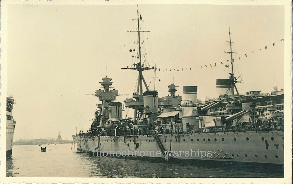 A photo of the two Trento-class heavy cruisers looking in 3/4 profile from astern. Behind are the distinctive striped funnels of the liner Conte di Savoia. In the distant background can be seen the shape of a Conte di Cavour-class battleship.