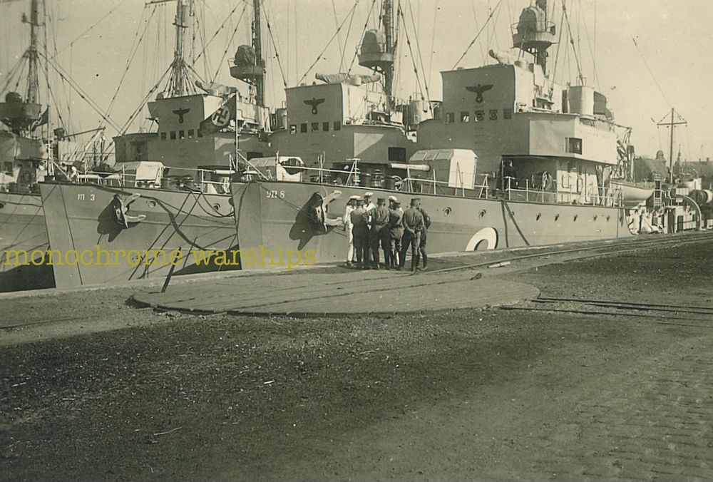 At least qty 3 Type 1935 M-class minesweepers of the Kriegsmarine docked alongside each other. Closest to quayside is M8, with M3 alongside it. A group of Kriegsmarine sailors are chatting on the quayside. In the foreground is a rail turntable, allowing trucks to be wheeled alongside.