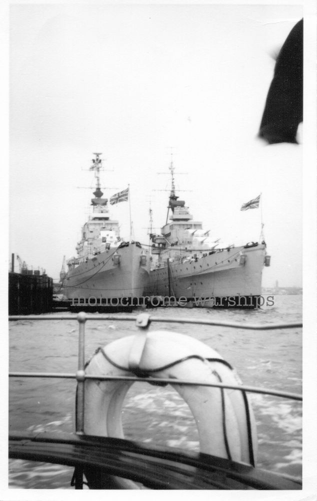 Qty 2 Dido / Bellona class AA cruisers moored alongside each other, dockside. The Union flag is flying proudly from both jackstaffs. We are looking bow on and can see qty 2 x 5.25" turrets on the left-hand ship, and qty 3 on the right. Photo appears to have been taken from a vessel on a harbour tour.