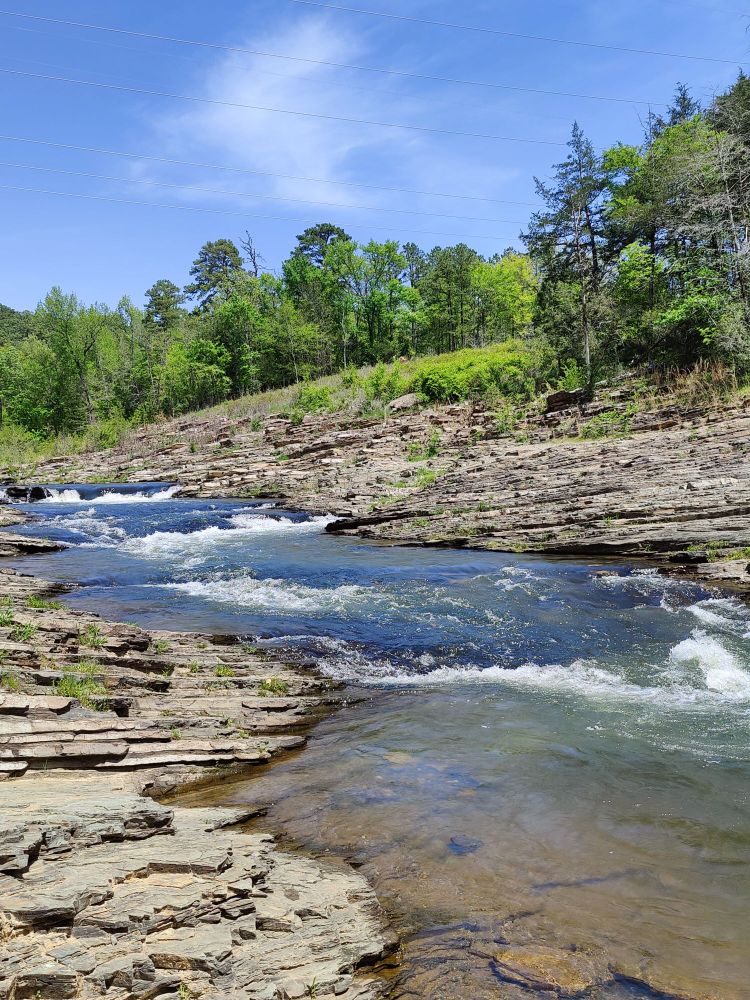 Clear, flowing water from the Mountain Fork river at Beaver Bend State Park. Alongside the river are walls of rock. Trees are visible in the distance.