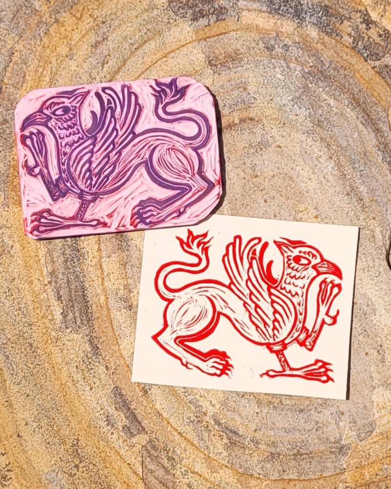 A pink linocut block (and resulting print, in red ink on brown paper) of a gryphon with a sly expression 