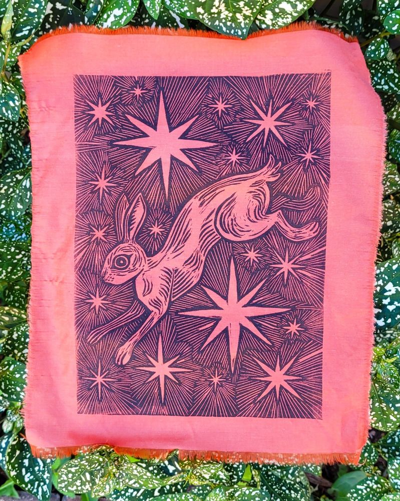 Block print in black ink on orange fabric of a rabbit jumping through stars 