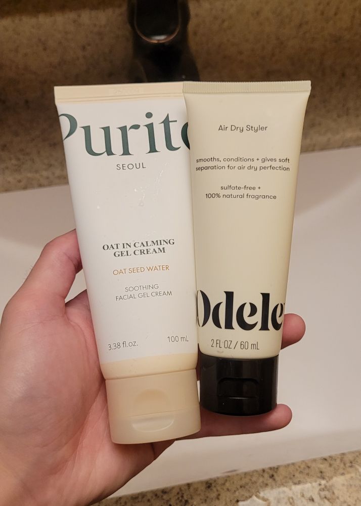two very similar cream-colored cosmetics bottles; one for purito oat face cream and one for odele air dry styler