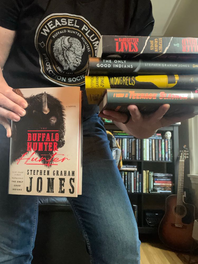 Me holding Buffalo Hunter Hunter in one hand and The Babysitter Lives, Killer on the Road, the Only Good Indians, Mongrels, I Was A Teenage Slasher